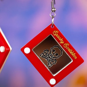 May include: Red plastic earrings shaped like a red Etch A Sketch toy with a white frame and a silver skull drawing on the screen. The text "Spooky Scribbler" is written in gold on the side of the earring.
