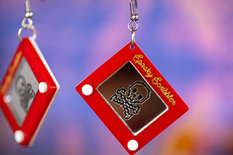 May include: Red plastic earrings shaped like a red Etch A Sketch toy with a white frame and a silver skull drawing on the screen. The text "Spooky Scribbler" is written in gold on the side of the earring.