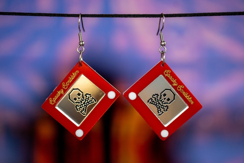 May include: A pair of red and silver earrings shaped like a classic red Etch A Sketch toy. The screen of each earring displays a pixelated skull and crossbones with the text "Spooky Scribbler" in gold lettering.
