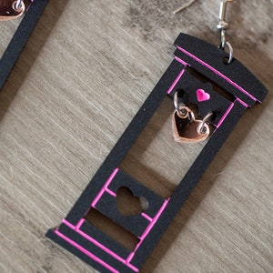 May include: A pair of black guillotine earrings with pink trim. Each earring features a small heart-shaped charm hanging from the blade.
