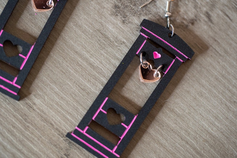 May include: A pair of black guillotine earrings with pink trim. Each earring features a small heart-shaped charm hanging from the blade.