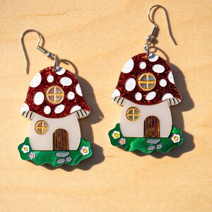 May include: A pair of red and white mushroom house earrings with green grass and white flowers. The mushroom houses have white walls, brown doors, and gold windows.