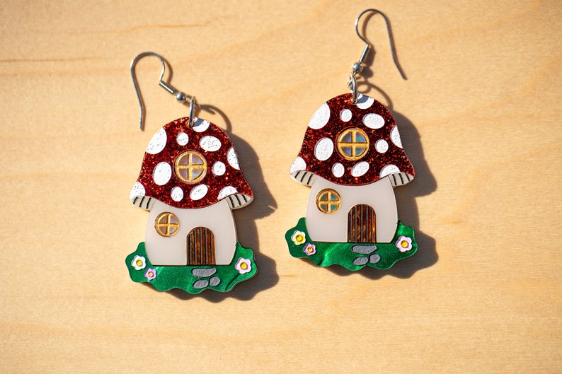 May include: A pair of red and white mushroom house earrings with green grass and white flowers. The mushroom houses have white walls, brown doors, and gold windows.