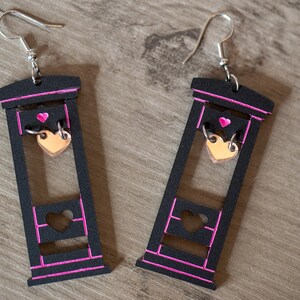 May include: A pair of black wooden earrings shaped like a guillotine with a pink outline. Each earring has a small gold heart dangling from the blade.
