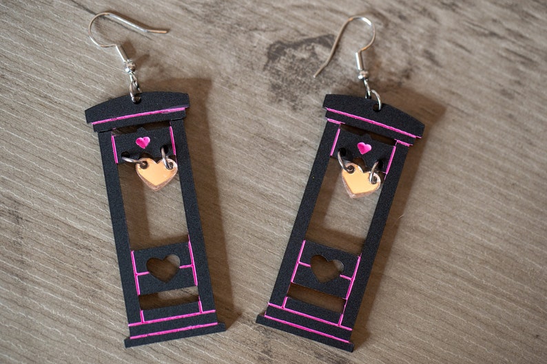 May include: A pair of black wooden earrings shaped like a guillotine with a pink outline. Each earring has a small gold heart dangling from the blade.