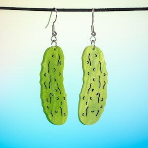 Peut inclure: Une paire de boucles d'oreilles en forme de cornichon vert avec des contours noirs. Les boucles d'oreilles sont suspendues à des crochets argentés.