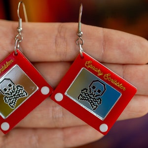May include: A pair of red earrings shaped like a classic red Etch A Sketch toy with a silver skull and crossbones design inside the drawing area. The earrings are labeled "Spooky Scribbler".