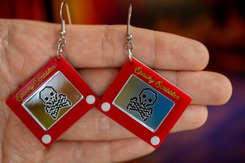 May include: A pair of red earrings shaped like a classic red Etch A Sketch toy with a silver skull and crossbones design inside the drawing area. The earrings are labeled "Spooky Scribbler".