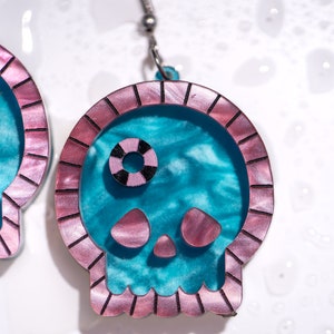 May include: A pair of skull-shaped earrings. The earrings feature a teal interior with a pink border and accents. The skull's eye is a black and white circle. The earrings are suspended from silver-colored hooks.