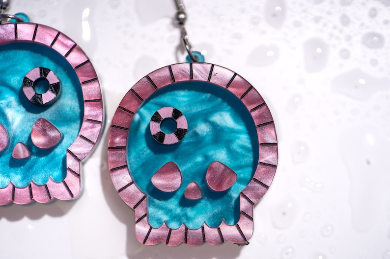 May include: A pair of skull-shaped earrings. The earrings feature a teal interior with a pink border and accents. The skull's eye is a black and white circle. The earrings are suspended from silver-colored hooks.