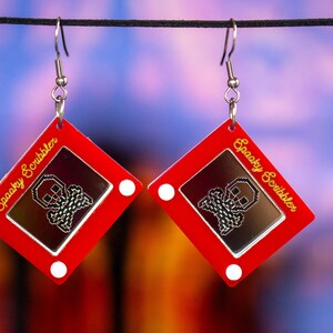 May include: A pair of red earrings shaped like a red Etch A Sketch toy with a silver skull drawn on the screen. The earrings have silver hooks and the text "Spooky Scribbler" is written in gold on the side of each earring.