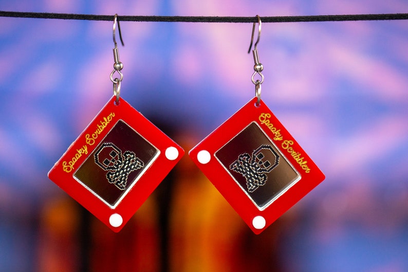 May include: A pair of red earrings shaped like a red Etch A Sketch toy with a silver skull drawn on the screen. The earrings have silver hooks and the text "Spooky Scribbler" is written in gold on the side of each earring.