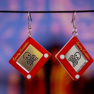 May include: A pair of red and silver earrings shaped like a red Etch A Sketch toy with a gold skull and crossbones design on the screen. The text "Spooky Scribbler" is written on the side of the toy.