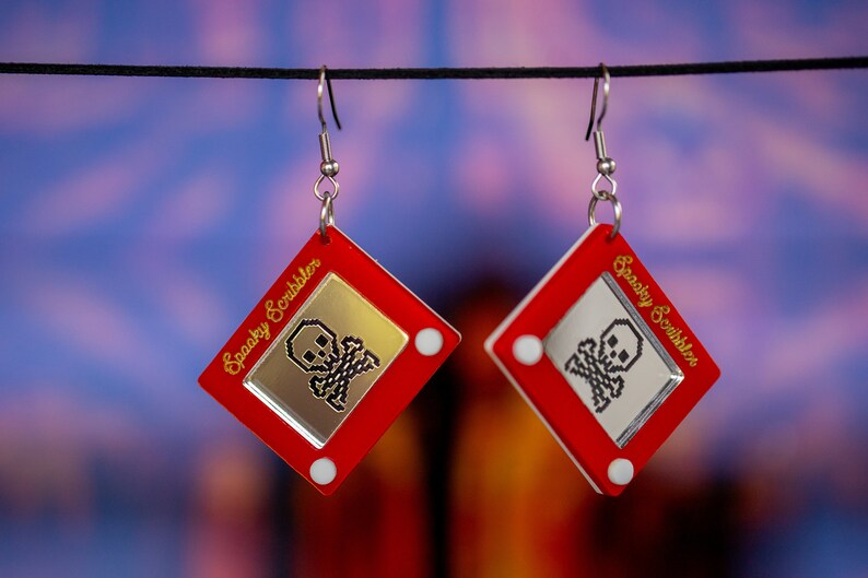 May include: A pair of red and silver earrings shaped like a red Etch A Sketch toy with a gold skull and crossbones design on the screen. The text "Spooky Scribbler" is written on the side of the toy.