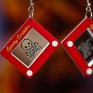 May include: A pair of red earrings shaped like a classic red toy drawing pad with a white frame. One earring has a pixelated skull and crossbones drawing and the text "Spooky Scribbler" on the frame. The other earring has a pixelated drawing of a ghost and the text "Spooky Scribbler" on the frame.