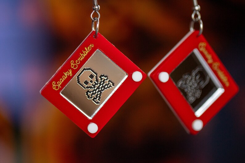 May include: A pair of red earrings shaped like a classic red toy drawing pad with a white frame. One earring has a pixelated skull and crossbones drawing and the text "Spooky Scribbler" on the frame. The other earring has a pixelated drawing of a ghost and the text "Spooky Scribbler" on the frame.
