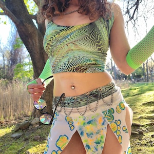 Könnte beinhalten: Ein grünes und gelbes Oberteil mit Muster und ein weißer Rock mit blauen und gelben Akzenten. Der Rock hat einen Kettenriemen und ein einzigartiges asymmetrisches Design. Das Outfit wird mit neongrünen Netzarmstulpen und runden Sonnenbrillen ergänzt.