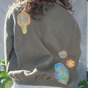 May include: An olive green bomber jacket adorned with vibrant, abstract patches. The patches showcase psychedelic designs in blue, orange, and yellow. The jacket features a ribbed hem and cuffs.