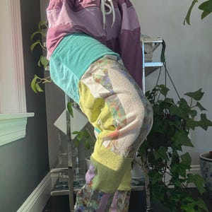Könnte beinhalten: Bunte Patchwork-Jogginghose in Lila-, Türkis-, Gelb- und Blumenmustern. Die Hose hat einen Kordelzug in der Taille und eine lockere Passform. Das Design zeichnet sich durch eine Mischung aus Stoffen und Texturen aus.