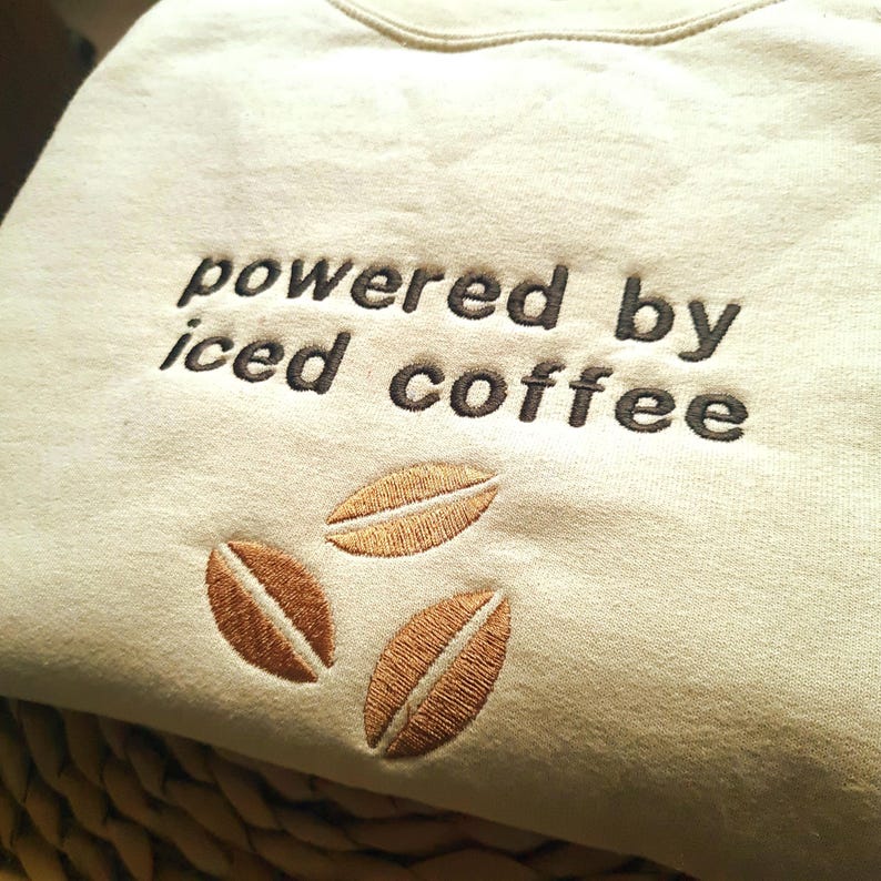May include: Beige sweatshirt with the embroidered text "powered by iced coffee" in dark brown. Three embroidered coffee bean designs are below the text, adding a coffee theme to the apparel.