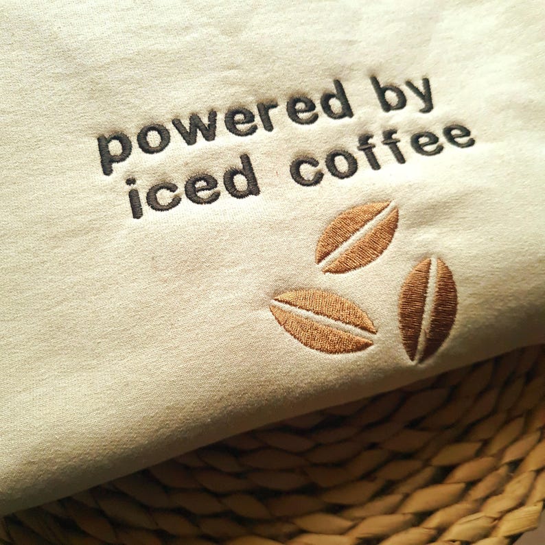 May include: Cream-colored fabric with the embroidered phrase "powered by iced coffee" in dark brown. Three embroidered coffee beans in shades of brown are below the text. The fabric is resting on a woven surface.