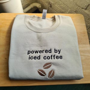 May include: A beige sweatshirt with the embroidered phrase "powered by iced coffee" and three brown coffee bean designs. The sweatshirt is folded neatly on a wooden surface.