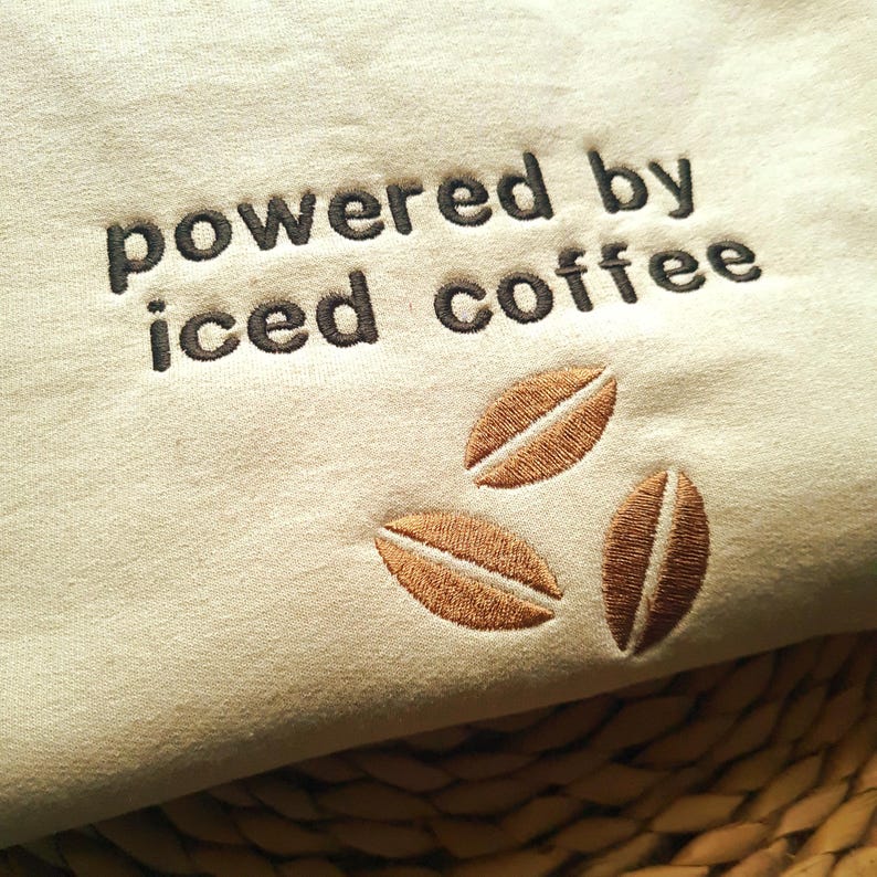 May include: Cream-colored fabric with the embroidered phrase "powered by iced coffee" in dark brown thread. Three brown coffee bean designs are embroidered below the text, creating a coffee-themed design.