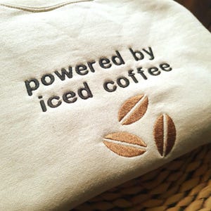 May include: Beige sweatshirt with the embroidered text "powered by iced coffee" in dark brown. Three coffee bean designs are embroidered in a lighter brown color.