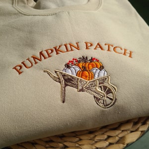 Könnte beinhalten: Ein beiger Pullover mit dem gestickten Text "Pumpkin Patch" in Orange. Unter dem Text befindet sich ein gesticktes Bild einer Schubkarre voller Kürbisse.