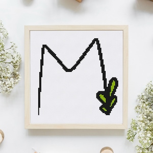 Könnte beinhalten: Ein gerahmtes Kreuzstich-Design des Buchstabens "M" mit einem grünen Blattdesign rechts neben dem Buchstaben.
