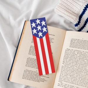 Puede incluir: Un marcapáginas de cuentas rojo, blanco y azul con un diseño de la bandera estadounidense. El marcapáginas está sobre un libro abierto.