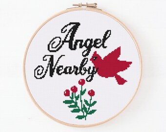May include: Cross stitch embroidery hoop with white fabric featuring the text "Angel Nearby" in black cursive font. A red cardinal bird with a black eye is flying above the text. A green plant with red berries is below the text.