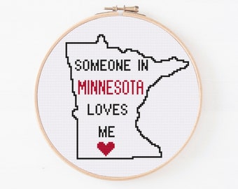 May include: A white embroidery hoop with a black outline of the state of Minnesota. The text "Someone in Minnesota loves me" is written in black with a red heart at the bottom.