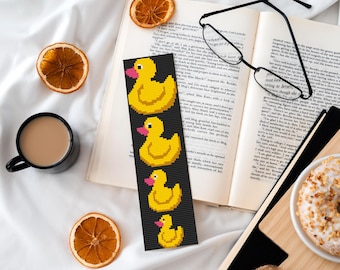 May include: A black bookmark with a row of four yellow rubber duckies with pink beaks. The duckies are different sizes and are arranged from largest to smallest. The bookmark is on top of an open book with a pair of reading glasses.