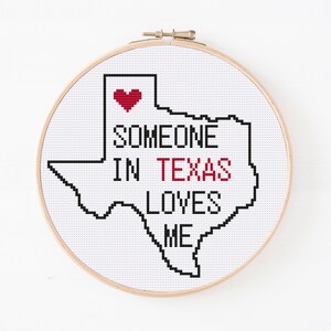 May include: A white cross-stitch embroidery hoop with a black outline of the state of Texas. Inside the outline, the text "Someone in Texas loves me" is written in black with a red heart above the word "Someone".