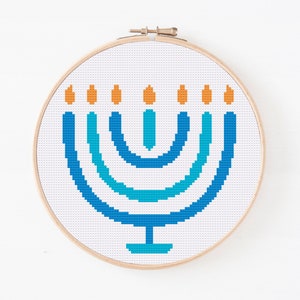 May include: A blue and turquoise menorah with orange flames on a white background. The menorah is in a wooden embroidery hoop.