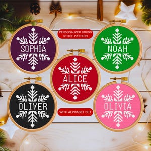 May include: Five round embroidery hoops with colorful backgrounds and white snowflake designs. Each hoop has a name in white letters: Sophia, Noah, Alice, Oliver, and Olivia. The text "Personalized Cross Stitch Pattern" and "With Alphabet Set" are also included.