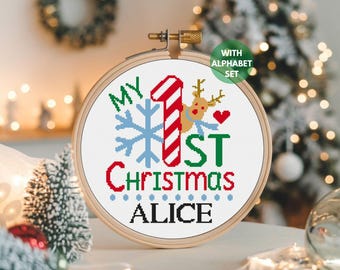 May include: A cross-stitch hoop art piece with the text "MY 1ST CHRISTMAS ALICE". The design includes a snowflake, a candy cane-style number one, a reindeer, and a heart. The text is in green, red, and blue. The hoop is in a wooden frame.