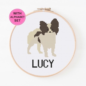 May include: Cross stitch pattern of a Papillon dog with the name Lucy underneath. The pattern is in a wooden embroidery hoop.