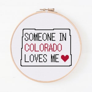 May include: Cross-stitch embroidery hoop with a white background and black text that reads "Someone in Colorado loves me" with a red heart.
