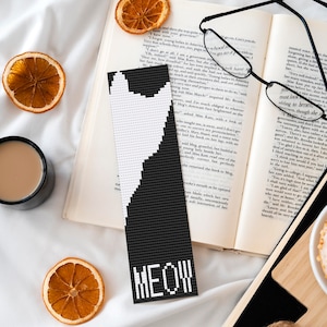 May include: A black and white cat bookmark with the word "MEOW" in white letters. The bookmark is lying on a book with a cup of coffee, dried orange slices, and a donut on a wooden cutting board.