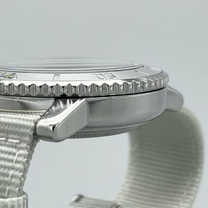 Puede incluir: Un reloj de pulsera plateado con una correa de nailon blanca. El reloj tiene un bisel giratorio con n&uacute;meros y marcas.
