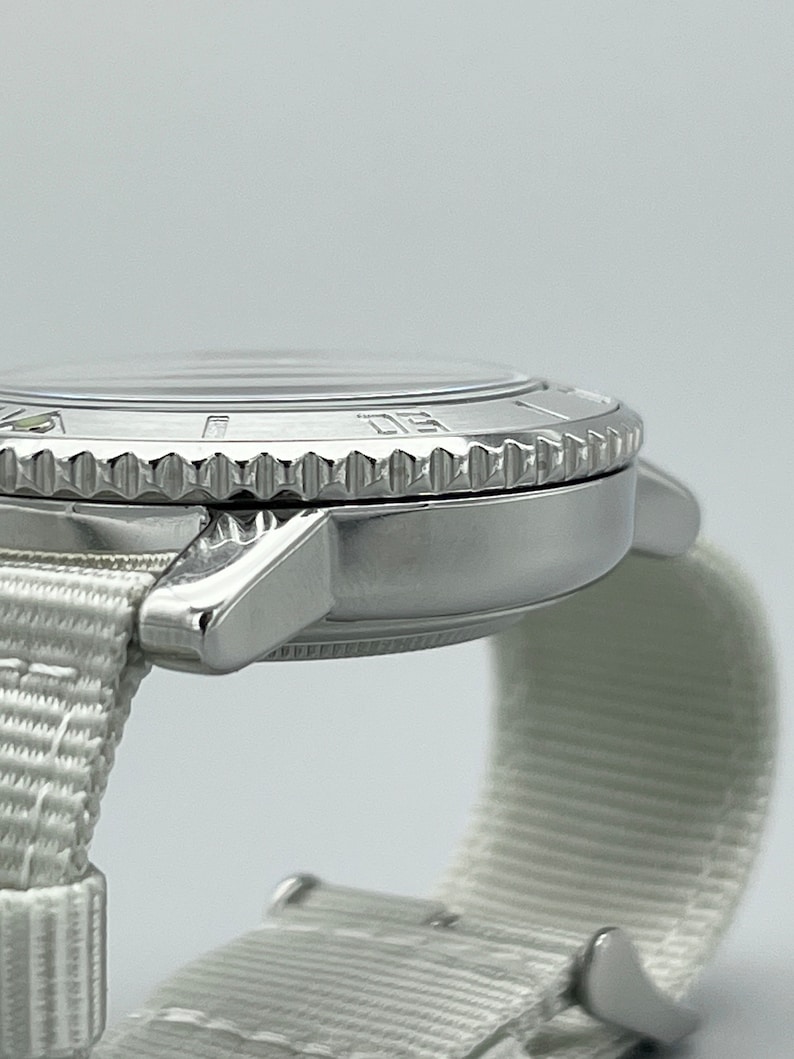 Puede incluir: Un reloj de pulsera plateado con una correa de nailon blanca. El reloj tiene un bisel giratorio con n&uacute;meros y marcas.