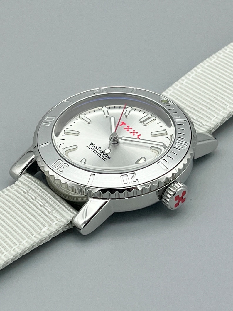 Puede incluir: Un reloj de pulsera plateado con una esfera blanca y detalles rojos. El reloj tiene un bisel negro con marcas blancas y una correa de nailon blanca.