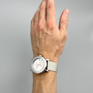 Puede incluir: Un reloj de pulsera plateado con una esfera blanca y una correa de nailon blanca. La esfera del reloj tiene marcas negras y una aguja de segundos roja.
