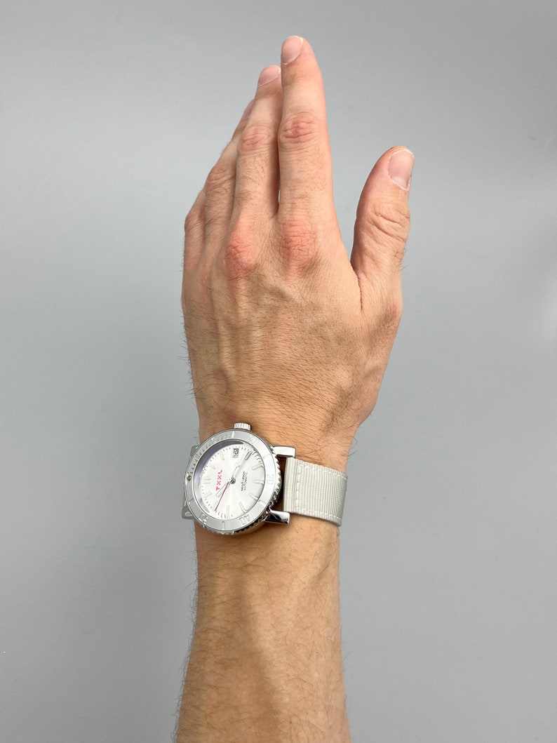 Puede incluir: Un reloj de pulsera plateado con una esfera blanca y una correa de nailon blanca. La esfera del reloj tiene marcas negras y una aguja de segundos roja.
