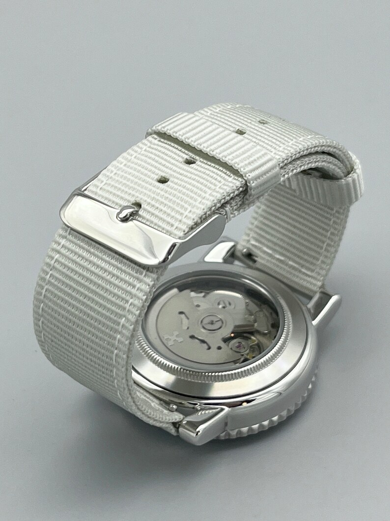 Puede incluir: Un reloj plateado con una correa de nailon blanca. La esfera del reloj es visible y muestra el funcionamiento interno del reloj.