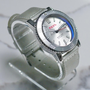 Puede incluir: Un reloj de pulsera plateado con una esfera blanca y una correa de nailon gris. La esfera del reloj tiene un logotipo "TXXXL" rojo y las palabras "660ft/200m AUTOMATIC".
