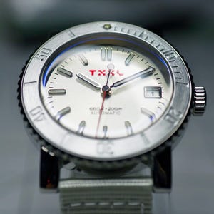 Puede incluir: Un reloj de pulsera plateado con una esfera blanca y detalles rojos. El reloj tiene un bisel negro con marcas blancas y las palabras "660ft - 200m AUTOMATIC" impresas en la esfera. El reloj tiene una correa de nailon blanca.