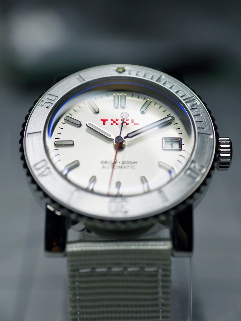 Puede incluir: Un reloj de pulsera plateado con una esfera blanca y detalles rojos. El reloj tiene un bisel negro con marcas blancas y las palabras "660ft - 200m AUTOMATIC" impresas en la esfera. El reloj tiene una correa de nailon blanca.